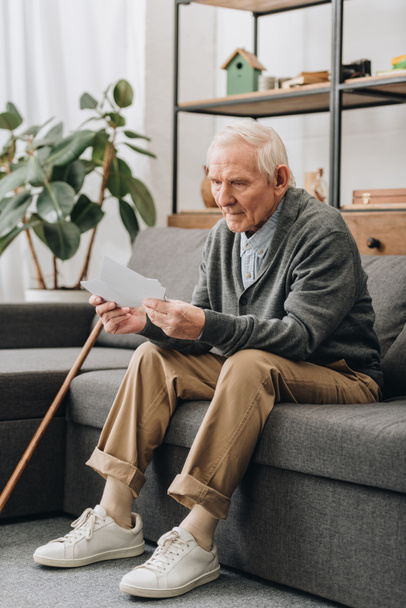 Trauriger Rentner mit grauen Haaren schaut sich Fotos an, während er auf dem Sofa sitzt  - Foto, Bild