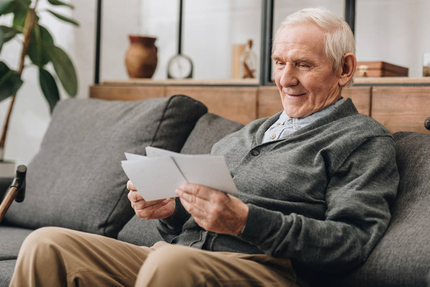 glücklicher älterer Mann mit grauen Haaren schaut sich Fotos an und sitzt auf dem Sofa - Foto, Bild