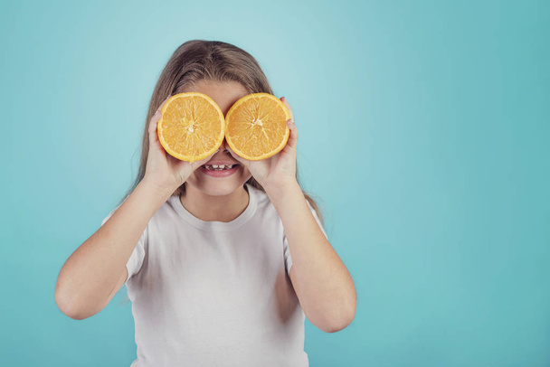 smiling little girl holding oranges over her eyes on blue background - Foto, afbeelding