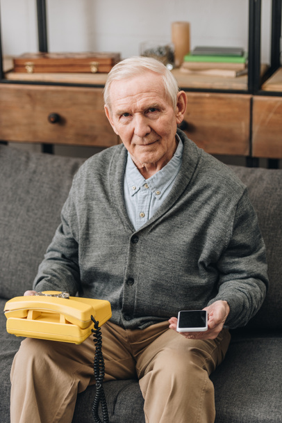 hombre retirado sosteniendo teléfono inteligente y teléfono retro mientras está sentado en el sofá
 - Foto, Imagen