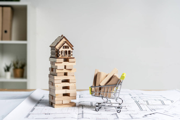 Little wooden house on top of jenga blocks with wood element or building plank board in small shopping cart stand on table inside bright workstation with copy space for text - Fotó, kép