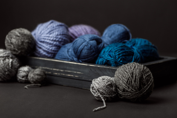 close up view of grey, purple and blue knitting clews in wooden box on black background - Фото, изображение