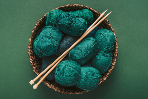 top view of green and blue yarn clews and knitting needles in wicker basket on green backdrop - Foto, Bild