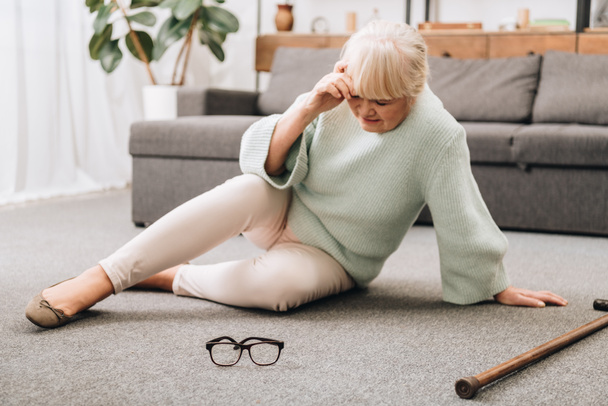 退職女性はメガネと歩行杖を有し頭痛の近くの床の上に座って  - 写真・画像