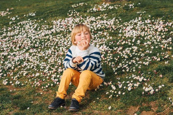 Весняний портрет чарівного маленького хлопчика, який відпочиває на траві, повний ромашкових квітів, в морському пуловері з смугою, жовті штани з чилі
 - Фото, зображення