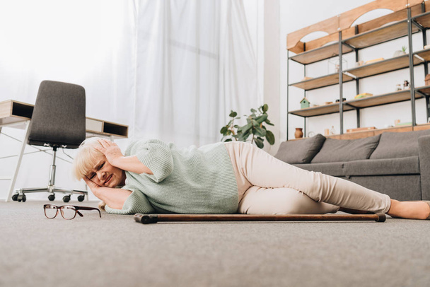 μοναχική συνταξιούχος γυναίκα ξαπλωμένη στο πάτωμα στο σαλόνι και κρατώντας το κεφάλι - Φωτογραφία, εικόνα