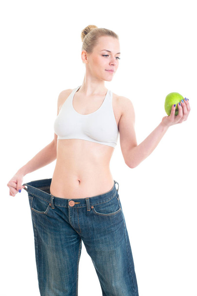 Het losmaken van gewicht vrouw in grote spijkerbroek duim opdagen. Slanke lichaam zorg meisje poseren in studio met groene appel. - Foto, afbeelding