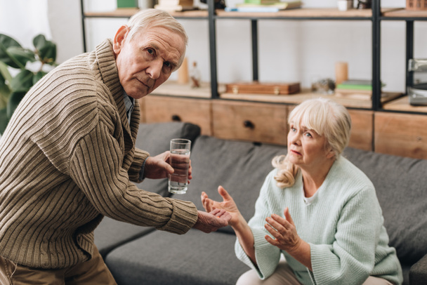 senior man giving to old woman glass of water and pills while looking at camera - Photo, Image