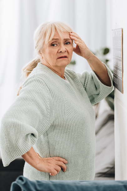 засмучена старша жінка стоїть біля календаря на стіні і тримає голову
 - Фото, зображення