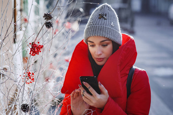 close up portrait of happy woman in red winter down jacket and knitted grey hat types text of sms or message  in her mobile smartphone standing on the winter street near traditional christmas tree.  Christmas, new year and winter holiday concept - Im - Fotografie, Obrázek