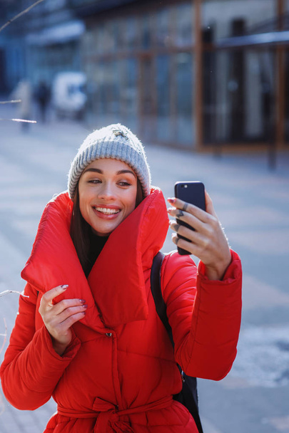 Winter selfie. Young happy girl in red down jacket and grey knitted hat takes selfie and play the ape in winter cold day. Girl takes winter selfie. Christmas, new year and winter holiday concept - Image - Фото, зображення