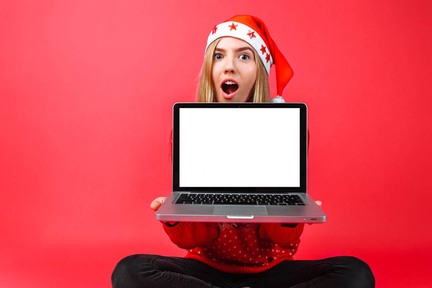 Excited by the good news woman, in Santa's hat, holding a blank laptop screen, copy space for text. Beautiful young smiling female model isolated on red background. - Fotografie, Obrázek