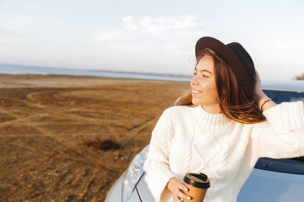 Beautiful young woman outdoors at the beach during sunset, sitting on a car, drinking coffee - Photo, image