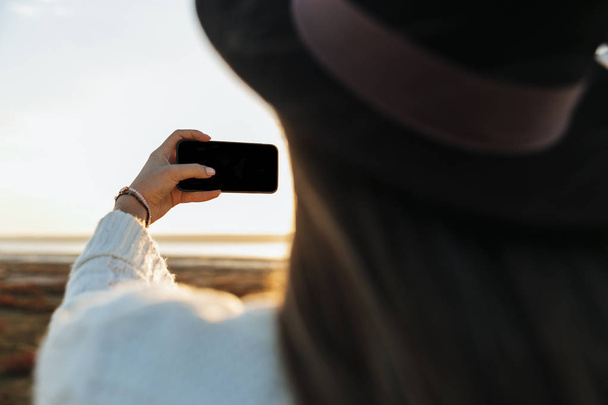 Rückansicht einer jungen Frau, die während des Sonnenuntergangs draußen am Strand spaziert und ein Selfie macht - Foto, Bild