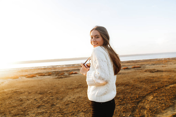 Beautiful young woman walking outdoors at the beach during sunset, taking pictures with photo camera - Foto, Bild