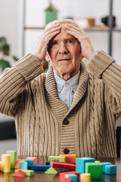 pensionista segurando cabeça enquanto sentado perto de brinquedos de madeira em casa
  - Foto, Imagem