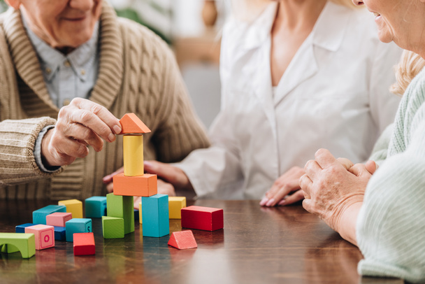 cropped view of caregiver sitting with retired man and woman and playing with wooden toys  - Foto, afbeelding