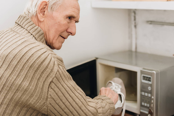 senior man with dementia disease putting shoe in microwave oven  - Фото, изображение