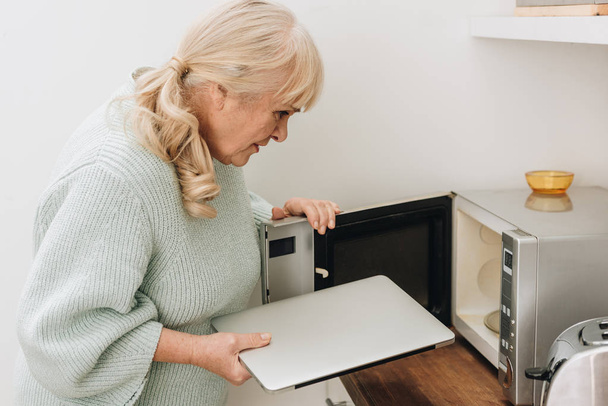 senior woman with dementia disease putting laptop in microwave oven  - Foto, afbeelding