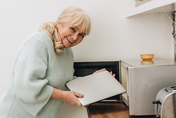 cheerful senior woman with dementia disease putting laptop in microwave oven - Fotó, kép