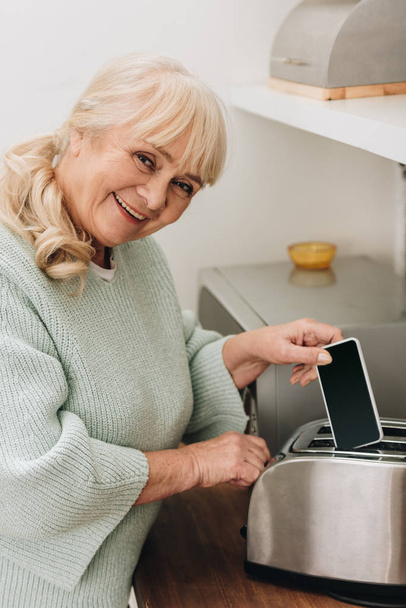 cheerful retired woman with alzheimer disease putting smartphone in toaster  - Photo, Image