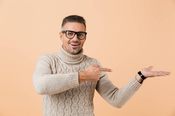 Portrait of a cheerful man dressed in sweater standing isolated over beige background, pointing away at copy space on his palm - Foto, afbeelding
