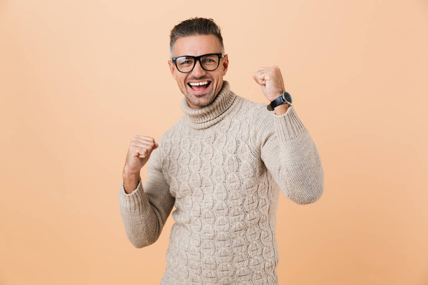 Portrait of a happy mature man dressed in sweater standing isolated over beige background, celebrating success - Foto, Imagen