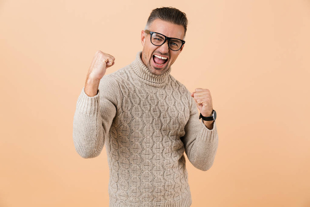 Portrait of a happy mature man dressed in sweater standing isolated over beige background, celebrating success - Fotoğraf, Görsel
