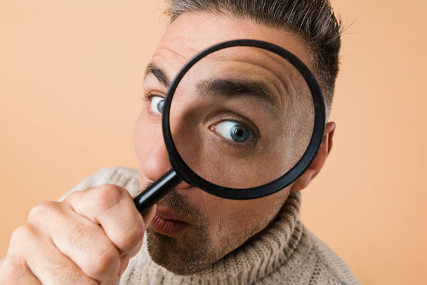 Close up of a man looking through a magnifying glass isolated over beige background - Fotografie, Obrázek