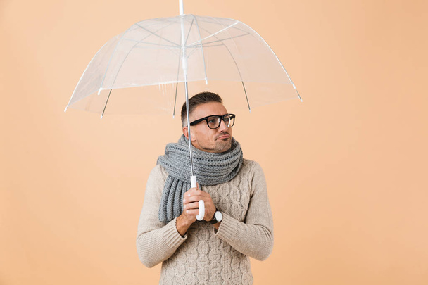 Portrait of a cold man dressed in sweater and scarf standing under umbrella isolated over beige background - Foto, imagen