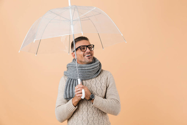 Portrait of a smiling man dressed in sweater and scarf standing under umbrella isolated over beige background - Фото, изображение