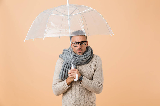 Portrait of a confused man dressed in sweater and scarf standing under umbrella isolated over beige background - Foto, afbeelding