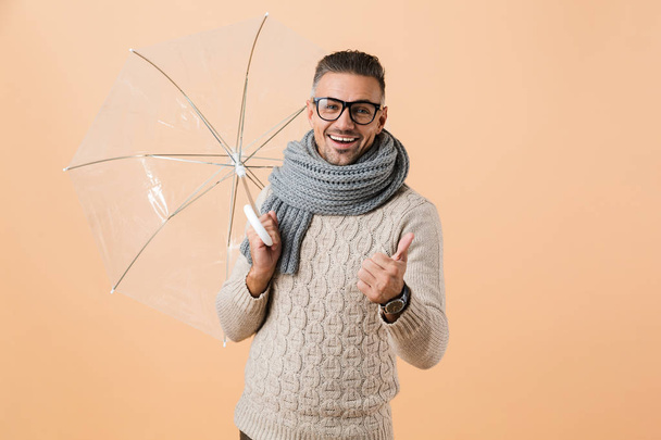 Portrait of a cheerful man dressed in sweater and scarf standing under umbrella isolated over beige background, showing thumbs up - Foto, imagen
