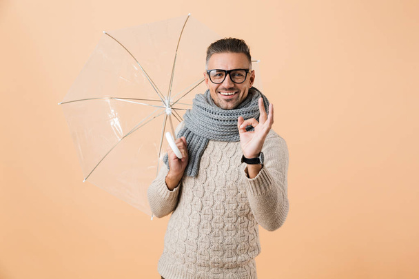 Portrait of a cheerful man dressed in sweater and scarf standing under umbrella isolated over beige background, showing ok - Photo, image