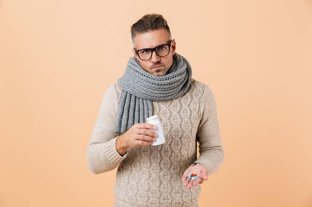 Portrait of an upset man dressed in sweater and scarf standing isolated over beige background, showing pills capsules - Fotografie, Obrázek