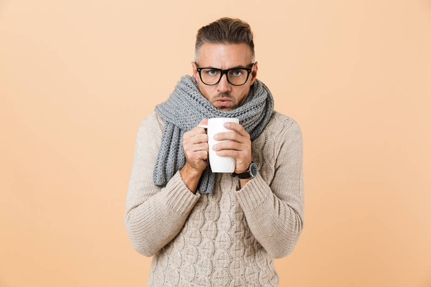 Portrait if a frozen man dressed in sweater and scarf holding cup of hot tea isolated over beige background - 写真・画像