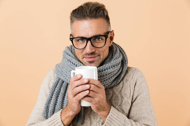 Portrait if a smiling man dressed in sweater and scarf holding cup of hot tea isolated over beige background - Photo, Image