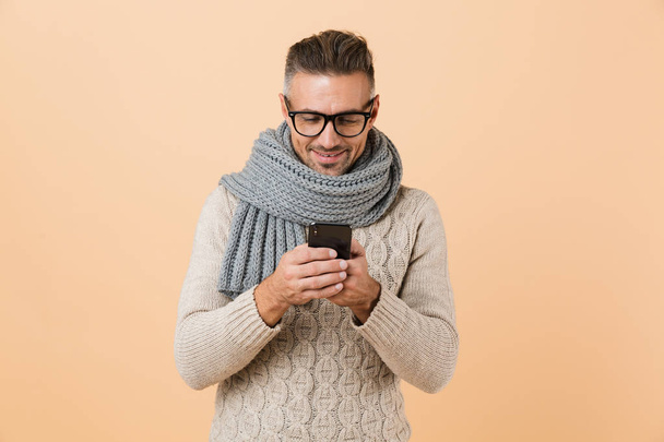 Portrait of a happy man dressed in sweater and scarf standing isolated over beige background, using mobile phone - Foto, Imagen