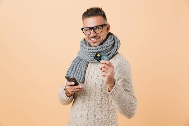Portrait if a happy man dressed in sweater and scarf standing isolated over beige background, holding mobile phone, showing credit card - Photo, Image