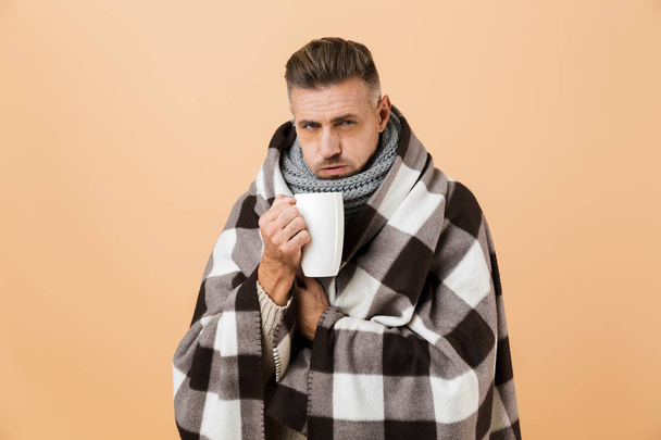Portrait of a sick man wrapped in a blanket standing isolated over beige background, holding cup with hot tea - Foto, imagen