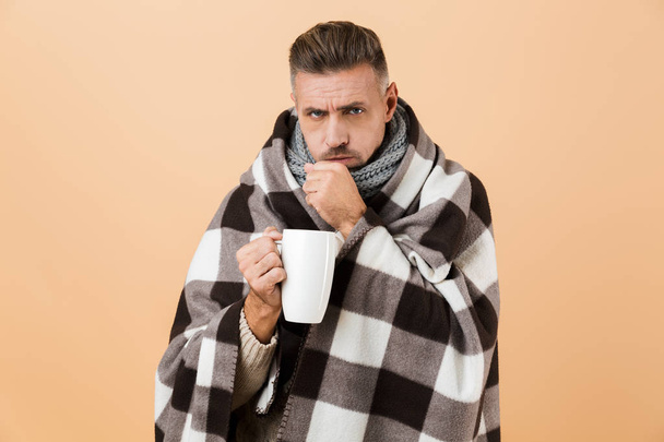 Portrait of a sick man wrapped in a blanket standing isolated over beige background, holding cup with hot tea - Photo, Image