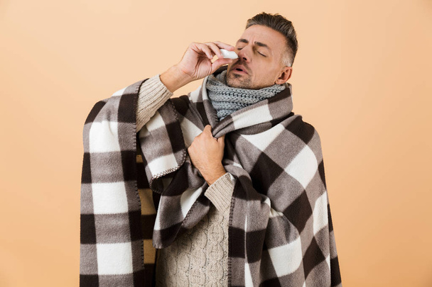 Portrait of a sick man wrapped in a blanket standing isolated over beige background, having runny nose, using napkin - Фото, зображення