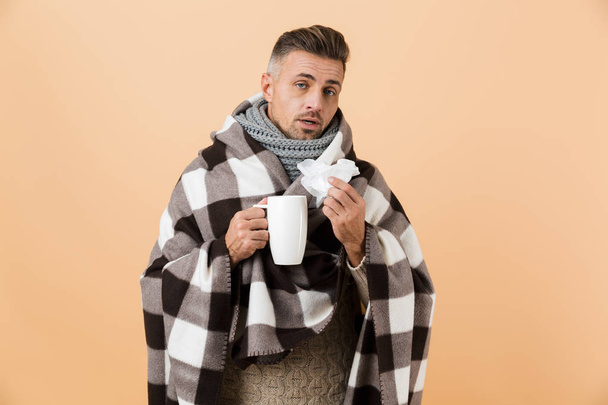 Portrait of a sad sick man wrapped in a blanket standing isolated over beige background, holding a cup and a napkin - Fotografie, Obrázek