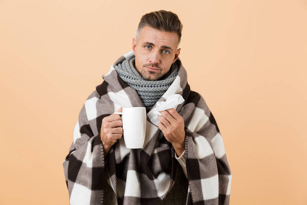 Portrait of a sick man wrapped in a blanket standing isolated over beige background, holding a cup and a napkin - Fotoğraf, Görsel