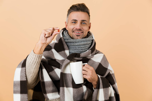 Close up portrait of a smiling man wrapped in a blanket standing isolated over beige background, showing pill capsule - Fotoğraf, Görsel
