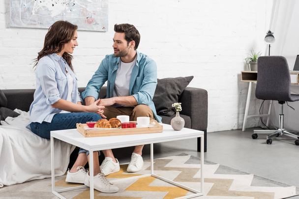 belo casal sentado no sofá e de mãos dadas enquanto toma café da manhã em casa
  - Foto, Imagem