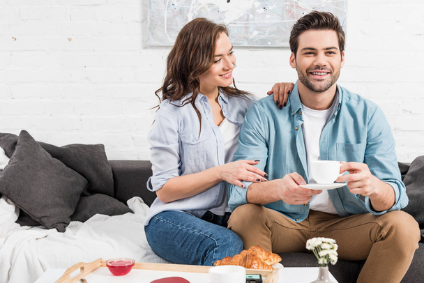 woman sitting on couch while smiling man drinking coffee during breakfast at home  - Foto, afbeelding