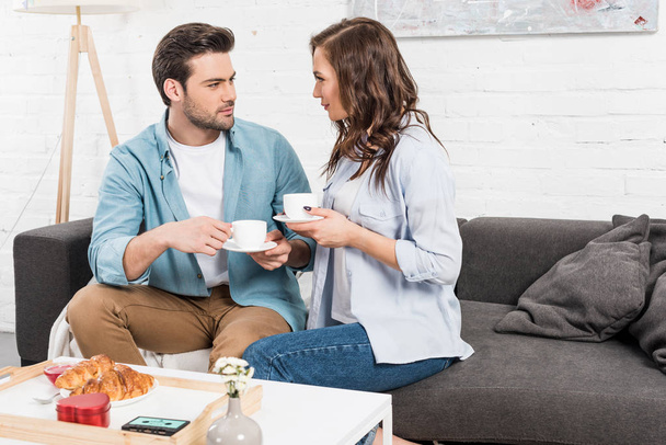 couple sitting on couch, looking at each other and drinking coffee during breakfast at home - Foto, Imagem