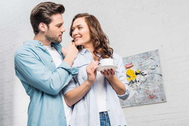man tenderly embracing attractive woman holding cup of coffee at home  - Fotoğraf, Görsel