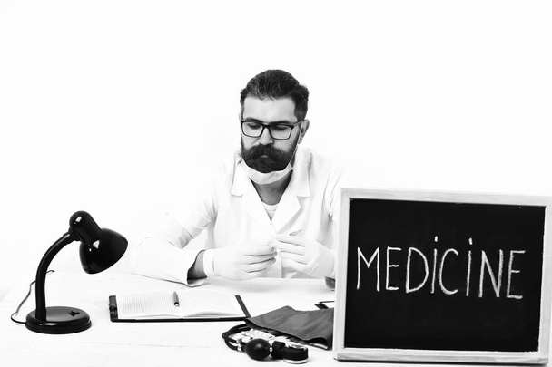 Bearded man, long beard. Brutal caucasian doctor or unshaven hipster in mask, medical gown sitting at table with board Medicine inscription isolated on white studio background. Medicine concep - Photo, Image